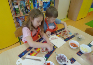 Dwójka dzieci dekoruje szynką, warzywami i serem placek w kształcie koła.